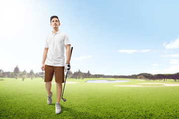 Handsome asian man in white clothes holding iron golf club walking on the golf course with sand bunkers, pond and trees