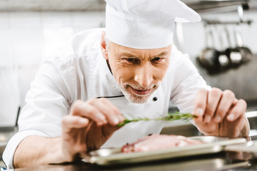 male chef in uniform decorating dish with herb in restaurant kitchen