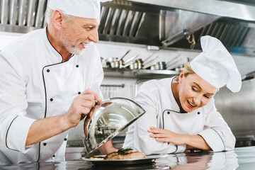 male chef in uniform presenting meat dish on serving tray to female cook in restaurant kitchen