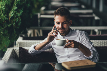 Handsome young business man, talking on the phone and smiling while a enjoying the cup of coffee in cafe