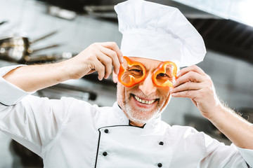 smiling male chef in uniform and hat holding pepper slices in front of face in restaurant kitchen