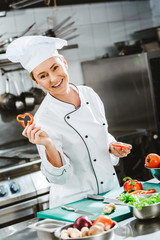 smiling female chef in uniform holding slices of pepper and looking at camera while cooking in restaurant kitchen