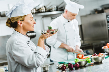 female chef in uniform holding meat dish on plate with male colleague cooking on background in...
