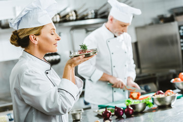 female chef in uniform holding meat dish on plate with male colleague cooking on background in restaurant kitchen