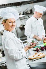 female chef in uniform holding meat dish on plate with male colleague cooking on background in restaurant kitchen