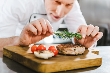 close up of male chef in uniform decorating meat steak with rosemary in restaurant kitchen