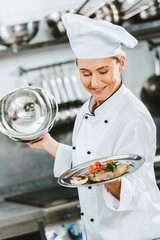 selective focus of beautiful female chef in uniform holding serving tray with meat dish in restaurant kitchen