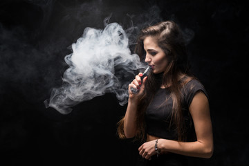 Attractive trendy woman in black shirt vaping and blowing smoke, isolated on dark background