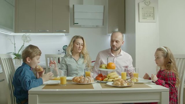 Pleasant positive family with lovely elementary age siblings having morning meal together in domestic kitchen. Cheerful casual parents and children enjoying breakfast while sitting at kitchen table.