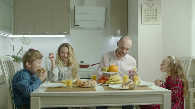 Joyful carefree family with two elementary age children having fun, staining noses with cream and laughing while enjoying healthy morning breakfast in domestic kitchen during weekend.