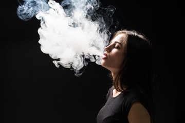 Young woman vaping and blowing smoke of electronic cigarette isolated on black background