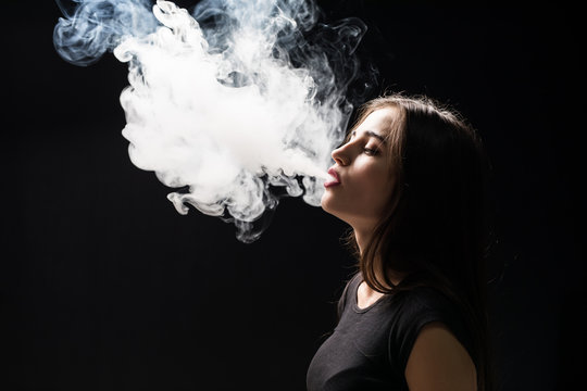 Young woman vaping and blowing smoke of electronic cigarette isolated on  black background Stock Photo | Adobe Stock