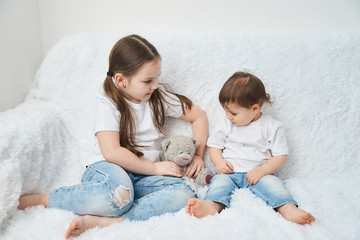 Two children, sisters sit on a white sofa in white t-shirts and blue jeans. Soft plush bear
