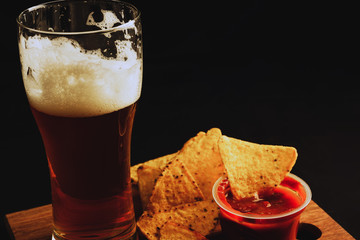 A glass of beer on a black background, nachos chips with sauce in the background.