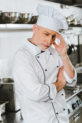 tired male chef in uniform touching head and having headache in restaurant kitchen