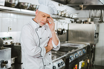 tired male chef in uniform touching head and having headache in restaurant kitchen