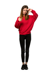 A full-length shot of a Young woman with red sweater making good-bad sign. Undecided between yes or not over isolated white background