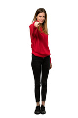 A full-length shot of a Young woman with red sweater points finger at you with a confident expression over isolated white background