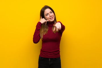 Woman with turtleneck over yellow wall making phone gesture and pointing front