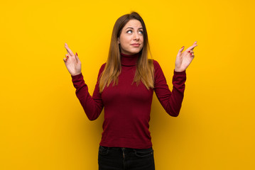 Woman with turtleneck over yellow wall with fingers crossing and wishing the best
