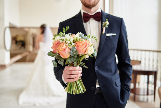 Close up of beautiful bridal bouquet of coral roses and greenery in groom's hand with bride in blur on background indoors, copy space. Happy wedding couple before ceremony