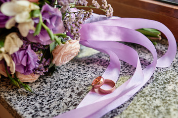 Close up of two golden wedding rings on purple satin ribbon of bridal bouquet in blur on marble window sill, copy space. Wedding concept
