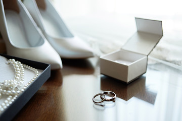 Luxury golden wedding rings with diamonds, white women shoes and bride jewelry (pearls necklace and earrings) on wooden background, copy space. Bridal accessories. Marriage concept. Close up