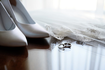 Beautiful golden wedding rings, white bride's shoes on high heels, veil on wooden table, copy space. Wedding morning preparation. Bridal accessories. Marriage concept. Close up