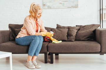 Senior woman in jeans cleaning sofa with spray and rag