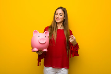 Young girl with red dress over yellow wall holding a piggybank