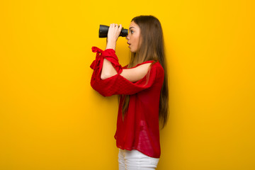 Young girl with red dress over yellow wall and looking in the distance with binoculars
