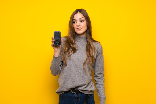 Young woman over yellow wall with troubled holding broken smartphone