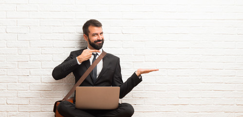 Businessman with his laptop sitting on the floor holding copyspace imaginary on the palm to insert...