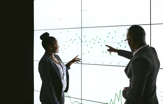 Silhouette Of A Black African Business People Giving A Presentation Conference. Pointing At A Large Video Screen With Charts And Graphs Next To Her In A Dark Room