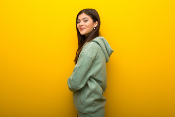 Obraz na płótnie Canvas Teenager girl with green sweatshirt on yellow background keeping the arms crossed in lateral position while smiling