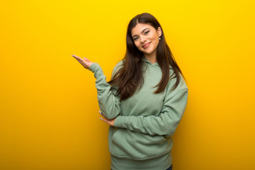 Teenager girl with green sweatshirt on yellow background extending hands to the side for inviting to come