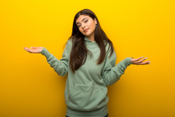 Teenager girl with green sweatshirt on yellow background making unimportant gesture while lifting the shoulders