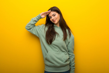 Teenager girl with green sweatshirt on yellow background saluting with hand
