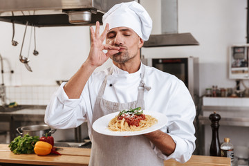 Photo of handsome male chief in white uniform holding plate with pasta, while cooking at kitchen in...