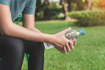 Young asian woman runner relaxing holding drinking water bottle and sitting on bench in the park outdoors after sport at early morning time, Exercise and healthy concept.