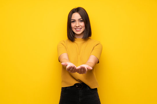 Young woman over yellow wall holding copyspace imaginary on the palm to insert an ad