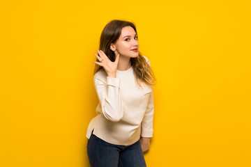 Teenager girl over yellow wall listening to something by putting hand on the ear