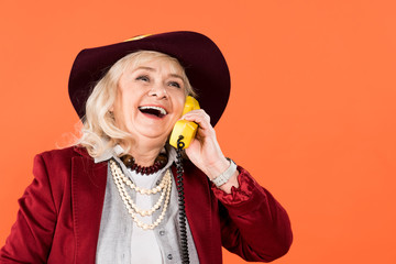 cheerful woman in hat talking on retro phone isolated on orange