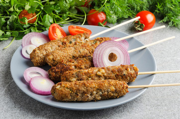 Closeup of served plate with kebab on wooden skewers,sliced onion,tomatoes,dill,cilantro on grey table
