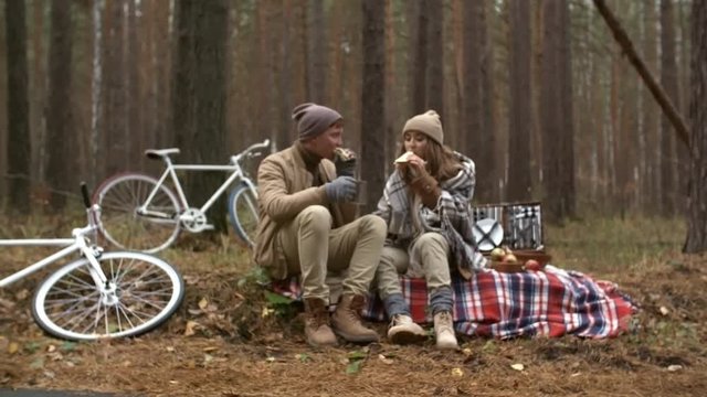 Zoom in of young boyfriend and girlfriend sitting on blanket beside bicycles in the forest and eating sandwiches while having picnic on autumn day
