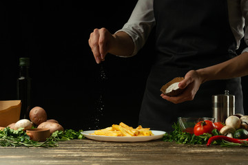 Chef salting french fries ready, on the background with vegetables. Cooking tasty but harmful food....
