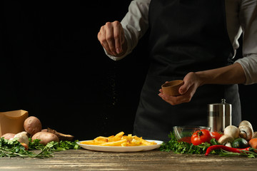The chef sprinkles ready-made french fries with herbs on the background with vegetables. Cooking tasty but harmful food. Background for design and menu, home-made recipe, cookbook. Fast food