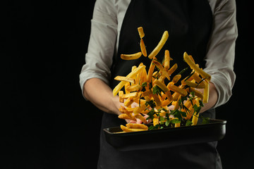 Professional chef prepares french fries with greens. Cooking tasty but harmful food on a dark background. Horizontal view.