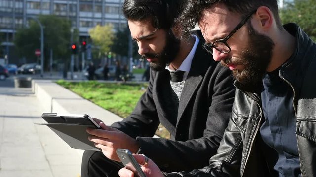 two young men outdoor using tablet and smart phone - remote working, multitasking, wireless technology concept