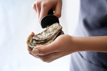 Woman opening raw oyster with knife against light background, closeup
